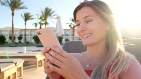 Portrait-of-female-resting-on-a-deckchair-using-smartphone,-browsing-the-internet,-scrolls-through-social-media.-Young-happy-girl-wearing-red-bikini-using-mobile-phone-while-sunbathing-by-the-pool