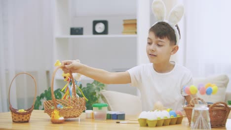 Cute-little-boy-with-bunny-ears-is-holding-a-wooden-basket,-full-of-Easter-colorful-eggs,-and-showing-with-his-finger-how-much-he-loves-Easter-conception.