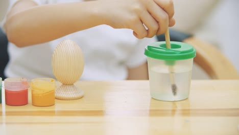 Creative-boy-is-mixing-and-stirring-water-in-a-jar-and-stirring-a-yellow-paint,-putting-in-a-paint-brush.