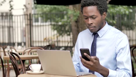 Afrikanischer-Geschäftsmann-nutzt-Smartphone-und-Laptop-im-Outdoor-Café