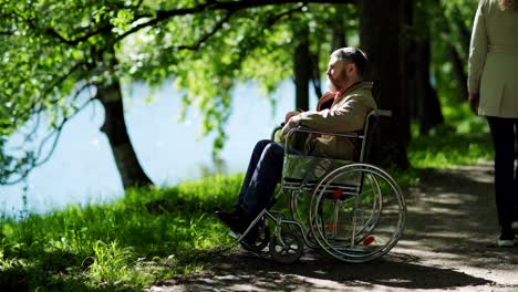 Inclinación-hacia-abajo-de-un-hombre-mayor-discapacitado-sentado-en-silla-de-ruedas-cerca-del-lago-en-el-parque-solo,-mirando-la-vista-y-pensando.-Mujer-irreconocible-pasando-por-aquí-en-un-día-ventoso