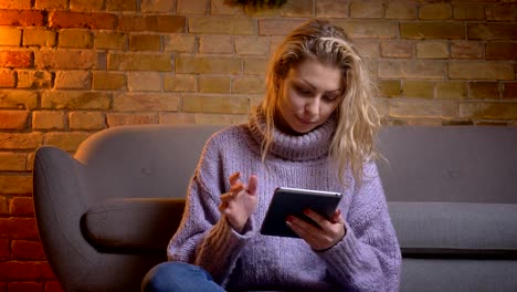 Closeup-top-view-shoot-of-adult-caucasian-blonde-female-using-the-tablet-looking-at-camera-and-laughing-while-sitting-on-the-floor-indoors-in-a-cozy-apartment