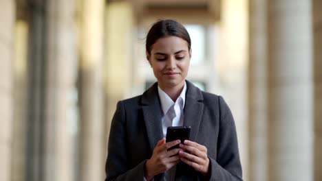 Cintura-arriba-tiro-de-atractiva-joven-en-abrigo-caminando-por-la-calle-después-del-trabajo.-Una-empresaria-sonriente-leyendo-mensajes-de-texto-en-el-teléfono-celular-o-navegando-por-las-redes-sociales