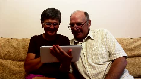 An-elderly-couple-is-sitting-on-a-sofa-at-home-and-watching-photos-on-a-tablet-pc,-smiling-and-talking-actively.