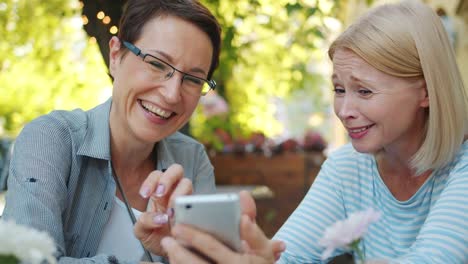 Weibliche-Freunde-mit-Smartphone-in-Straßencafé-Blick-auf-Bildschirm-Spaß-haben