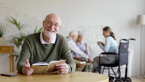 Enfoque-de-rack-de-tres-personas-mayores,-dos-mujeres,-incluyendo-discapacitados-uno-y-hombre,-jugando-a-las-cartas-en-el-hogar-de-ancianos.-Hombre-mayor-en-el-libro-de-lectura-de-gafas,-mirando-la-cámara-y-sonriendo-sentado-a-la-mesa