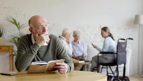 Rack-Fokus-von-drei-älteren-Menschen,-zwei-Frauen-einschließlich-behinderter-und-Mann,-Karten-im-Pflegeheim-spielen.-Intelligenter-Senior-in-Brille-lesen-Buch-und-denken-sitzen-am-Tisch