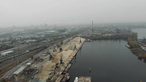 Port-cranes-unloading-river-sand-from-barge-in-docks-with-excavators-and-trucks-moving-around.-Industrial-docks-concept