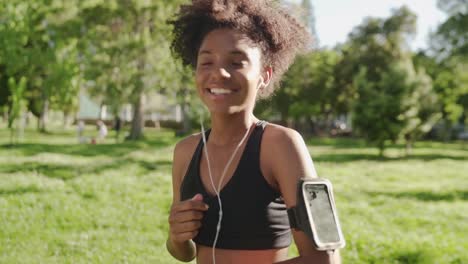 Happy-athlete-african-american-woman-runner-putting-earphones-in-her-ears-listening-to-music-on-smart-phone-in-armband-at-the-park