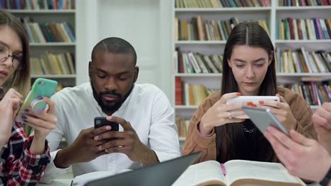 Vista-cercana-de-los-atractivos-estudiantes-multiétnicos-modernos-concentrados-que-utilizan-sus-teléfonos-durante-el-trabajo-sobre-la-tarea-universitaria-en-la-biblioteca