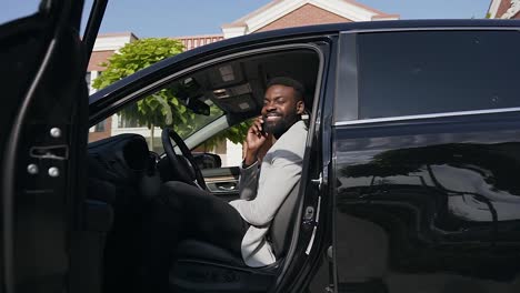 Joven-hombre-de-negocios-africano-en-traje-sentado-al-volante-de-coche-nuevo-en-el-aparcamiento-cerca-de-casa-mientras-se-habla-en-el-teléfono-inteligente.-Dolly-shot