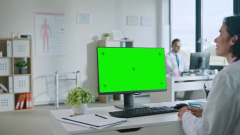 La-doctora-médica-está-haciendo-una-videollamada-con-el-paciente-en-un-ordenador-con-pantalla-de-simulación-de-pantalla-verde-en-una-clínica-de-salud.-Assistant-in-Lab-Coat-está-hablando-sobre-problemas-de-salud-en-la-oficina-del-hospital.