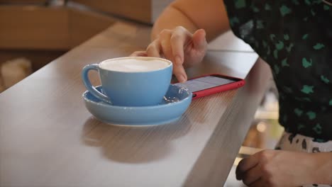 La-chica-está-escribiendo-en-el-teléfono-inteligente-mientras-está-sentada-en-una-cafetería.-Retrato-de-cerca-de-una-joven-feliz,-chica-sentada-en-un-café-bebiendo-café-por-la-mañana-en-la-ciudad.