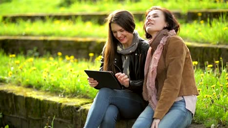 Junge-Frauen-mit-Toothy-Lächeln-sind-mittels-Tablet-in-Händen.-Attraktiven-Brünetten-sind-haben-Spaß-zusammen-mit-elektronischen-Gerät-im-Park-während-der-sonnigen-Frühling-Tag-im-freien