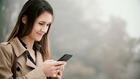 Portrait-of-young-beautiful-woman-in-foggy-park-in-the-morning.-Smiling-female-using-smartphone-with-touchscreen