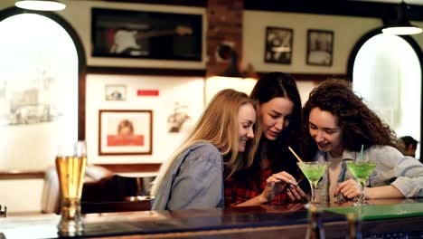 Las-niñas-son-con-smartphone,-riendo-y-hablando-mientras-estaba-sentado-en-la-barra-junto.-Chicas-en-ropa-casual-son-viendo-pantalla-y-charlando.-Tecnologías-modernas-para-el-concepto-de-la-diversión.