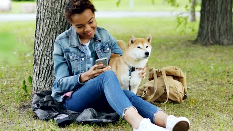 Happy-African-American-woman-is-using-smartphone-and-caressing-her-cute-pet-dog-resting-in-city-park-on-windy-summer-day.-Nature,-animals-and-people-concept.