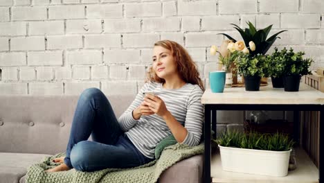 Attractive-girl-in-casual-clothing-is-using-smartphone-sitting-on-sofa-in-modern-loft-style-apartment-and-smiling.-Modern-technology,-interior-and-youth-concept.