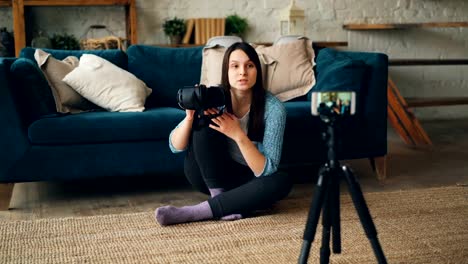 Bastante-joven-mujer-está-grabando-video-sobre-gafas-de-realidad-artificial-manteniendo-gadget-y-habla-mirando-a-smartphone-cámara-en-trípode-y-luego-agitando-la-mano.