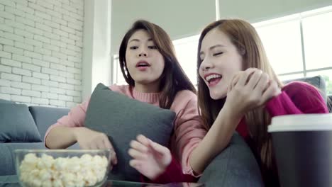 Asian-women-playing-pillow-fight-and-eating-popcorn-in-living-room-at-home,-group-of-roommate-friend-enjoy-funny-moment-while-lying-on-the-sofa.-Lifestyle-women-relax-at-home-concept.