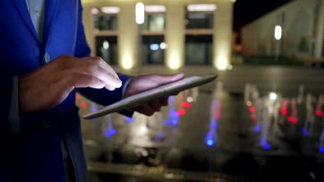 Hands-of-unrecognizable-businessman-using-digital-tablet-and-browsing-Internet-while-walking-down-night-city-street