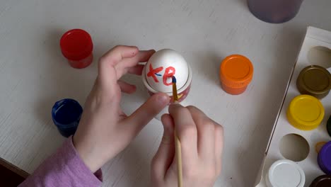 The-child-paints-Easter-egg-colors-and-draws-symbols-on-it-with-a-brush.-Close-up.-The-view-from-the-top.-4K.