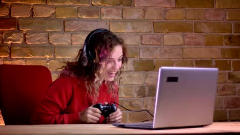 Portrait-of-young-female-blogger-in-red-hoodie-playing-video-game-using-joystick-and-winning-happily-on-bricken-wall-background.