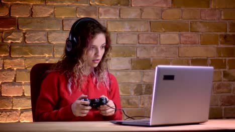 Portrait-of-young-female-blogger-in-red-hoodie-playing-video-game-using-joystick-and-failing-with-disgrace-on-bricken-wall-background.