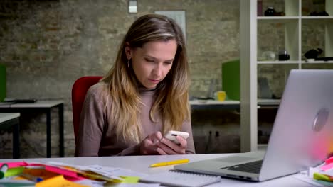 Closeup-of-attractive-caucasian-female-employee-using-phone-sitting-next-to-the-laptop-on-the-workplace