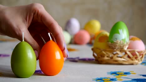 Weibliche-Hand-legt-auf-den-Tisch-drei-Osterkerzen.-Grün,-orange-und-gelb.-Bunte-Ostereier-im-Hintergrund.