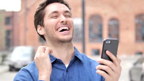 Outdoor-Portrait-of-Young-Man-Excited-for-Success-on-Smartphone