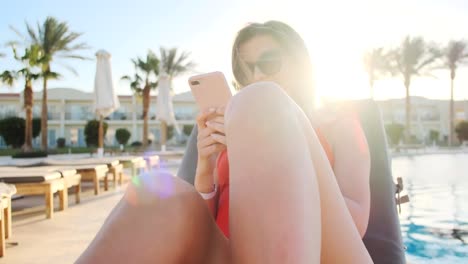 Young-girl-wearing-red-bikini-using-mobile-phone-while-sunbathing-by-the-pool.-Female-resting-on-a-deckchair-using-smart-phone,-browsing-the-internet,-scrolls-through-social-media.