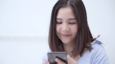 Joven-asiática-usando-teléfono-inteligente-mientras-se-acostó-en-la-cama-después-de-despertar-en-la-mañana,-hermosa-chica-japonesa-sonriente-relajarse-en-el-dormitorio-en-casa.-Disfrutar-del-tiempo-estilo-de-vida-mujeres-en-concepto-de-hogar.