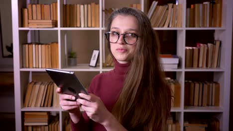 Nahaufnahme-Porträt-der-jungen-kaukasischen-Studentin-in-Brille-mit-dem-Tablet-lächelnd-und-Blick-auf-die-Kamera-drinnen-in-der-College-Bibliothek-drinnen