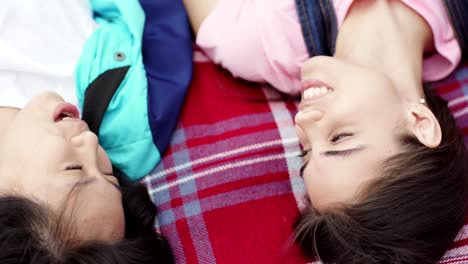 From-above-view-of-two-young-women,-Asian-and-Caucasian,-lying-on-plaid-blanket,-looking-at-each-other-and-talking,-closeup