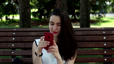 Retrato-de-cerca-Una-hermosa-joven-con-una-camiseta-blanca-está-chateando-en-las-redes-sociales-en-su-teléfono-inteligente-mientras-está-sentada-en-un-banco-en-un-parque.