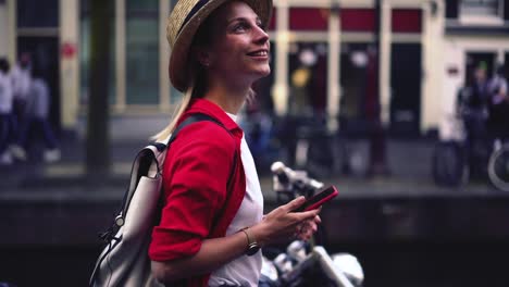 Side-view-of-smiling-Caucasian-hipster-girl-in-casual-wear-and-hat-walking-outdoors-on-city-street-and-holding-mobile-phone-in-hands