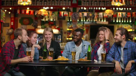 Afroamericano-con-amigos-en-el-bar-bebe-cerveza-y-come-patatas-fritas,-amigos-levantan-vasos-y-botellas-y-golpear-/-comprobar-en-la-mesa-sonriendo-y-diciendo-tostadas