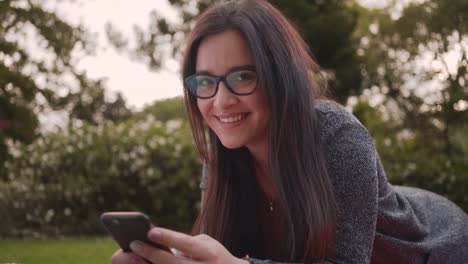 Retrato-de-una-hermosa-joven-con-anteojos-tumbados-en-el-parque-mirando-a-la-cámara-mientras-usa-sin-teléfono-inteligente-en-el-parque