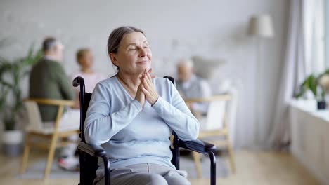 Tracking-shot-of-disabled-senior-woman-in-wheelchair-looking-away-and-contemplating-in-nursing-home,-other-aged-patients-in-background