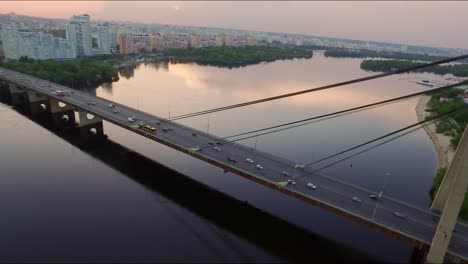 Luftbild-Auto-bewegt-sich-auf-Hängebrücke-über-Fluss.-Abendsonnenuntergang-in-der-Stadt