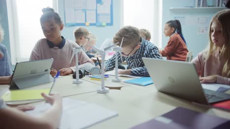 In-der-Grundschule:-Klasse-der-brillanten-kleinen-Kinder-arbeiten-als-Team-mit-Tablet-Computern,-um-Windkraftanlagen-zu-programmieren.-Klassenzimmer-mit-Kindern-Lernen-über-umweltfreundliche-Formen-erneuerbarer-Energie