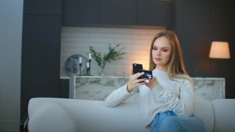 Una-mujer-hace-compras-en-línea-usando-un-teléfono-inteligente-y-una-tarjeta-de-banco-sentada-en-casa