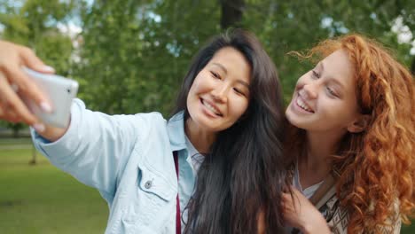 Chicas-lindas-tomando-selfie-con-el-teléfono-inteligente-posando-en-el-parque-verde-abrazando-sonriendo