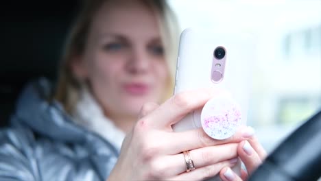 Blurred-blonde-woman-blogger-in-winter-clothes-sitting-in-car-and-talking-with-followers,-live-streaming,-looking-to-smartphone-screen.-focus-on-phone