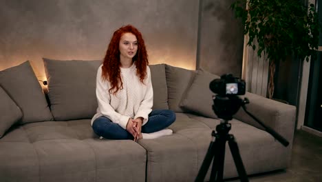 Attractive-curly-red-headed-young-girl-vlogger-is-talking-in-front-of-camera-recording-video-for-online-blog-in-internet-speaking,-smiling.-Woman-is-wearing-jeans-and-white-sweater