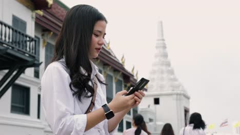 Young-Asian-female-using-a-smartphone-while-standing-on-the-street-beside-Wat-Phra-Kaew-in-Thailand-and-having-fun-in-vacation-time.