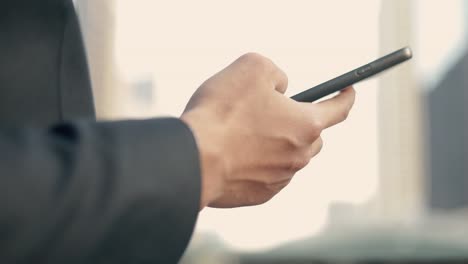 Close-up-a-businessman-in-a-black-suit-using-his-smartphone-browsing-financial,-scrolling-social-media-while-standing-outside-on-the-street-near-a-big-office-building-urban-having-sunlight-sunset.