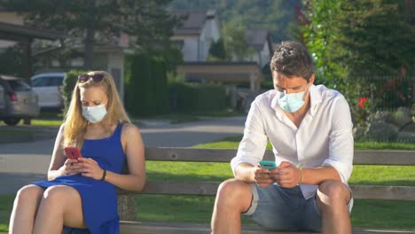 CLOSE-UP:-Two-strangers-wear-facemasks-while-sitting-on-park-bench-and-texting.