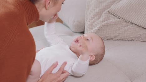 De-cerca-del-bebé-acostado-boca-arriba-en-el-sofá,-sonriendo-y-feliz,-estrechando-las-manos,-mirando-a-su-madre,-que-se-arrodilla-junto-al-bebé,-habla-con-él,-hace-cosquillas-y-sonríe